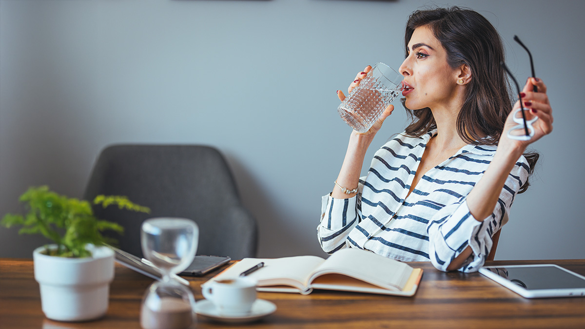 A young businesswoman taking off her glasses and drinking a big glass of water sitting behind her office desk